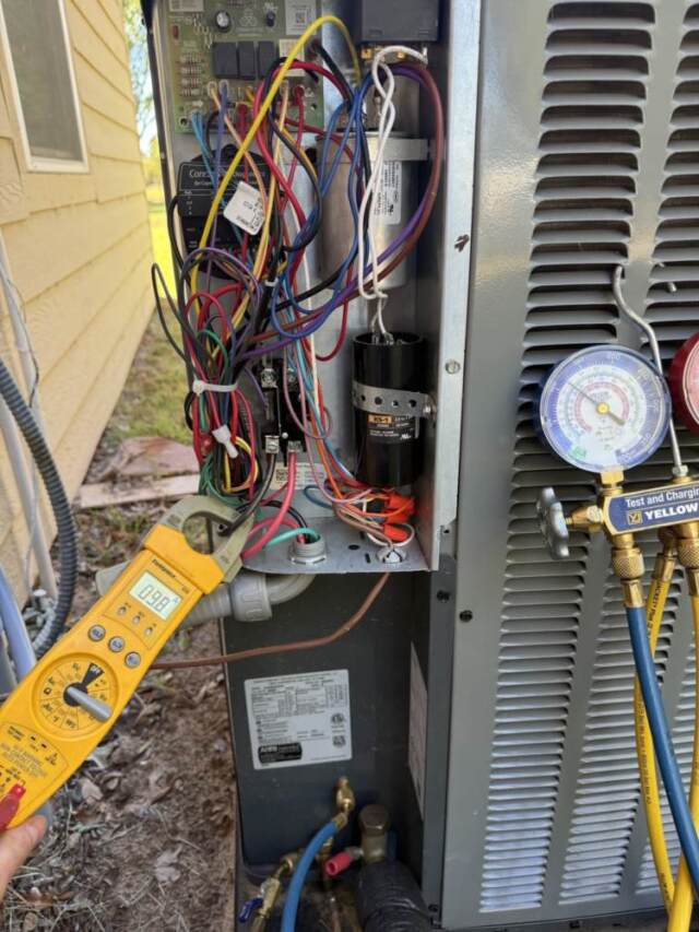 The cover is off the outdoor heat pump and an electrical meter is around a wire and the refrigerant guages are connected.
